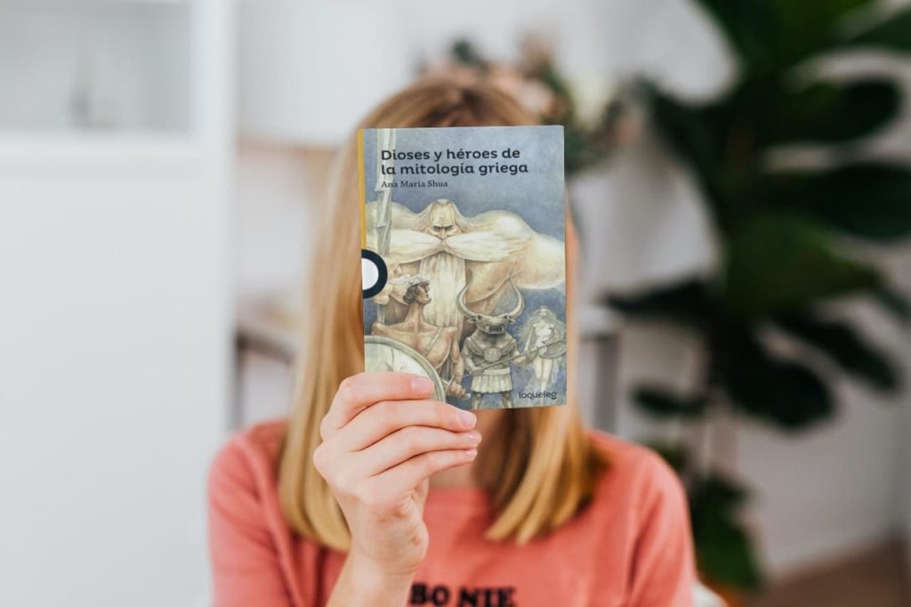 Persona cubriendo su rostro con el libro 'Dioses y héroes de la mitología griega' de Ana María Shua en un entorno interior con decoración de plantas