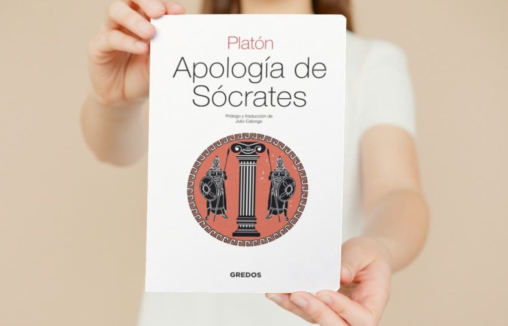 Una persona sosteniendo un libro titulado 'Apología de Sócrates' de Platón, con un diseño de portada que incluye columnas griegas y figuras de guerreros en un círculo rojo, publicado por Gredos.